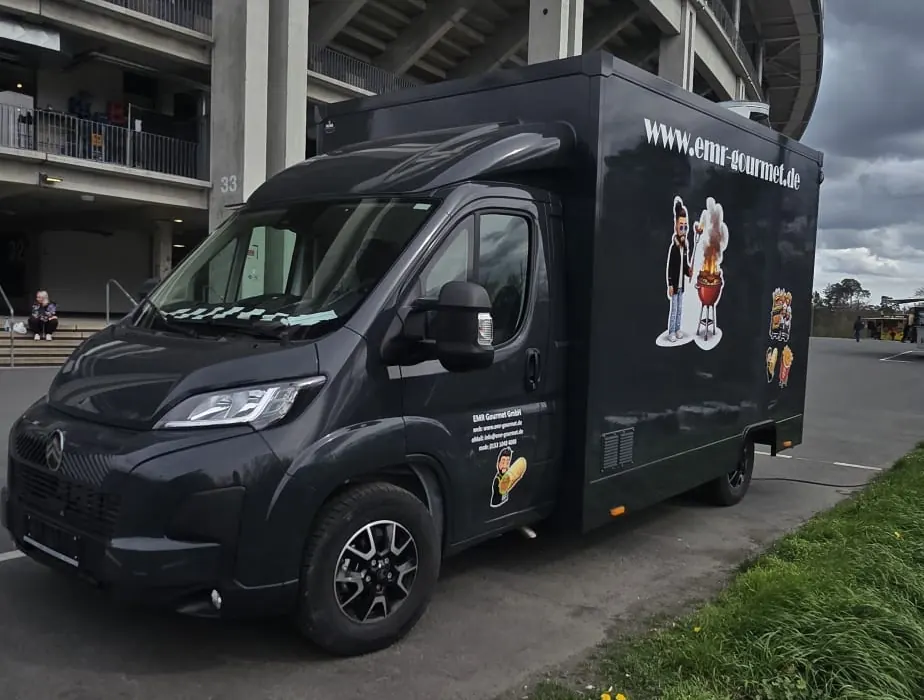 Der schwarze EMR Gourmet Foodtruck mit Logo und Webadresse, geparkt vor einem Stadion