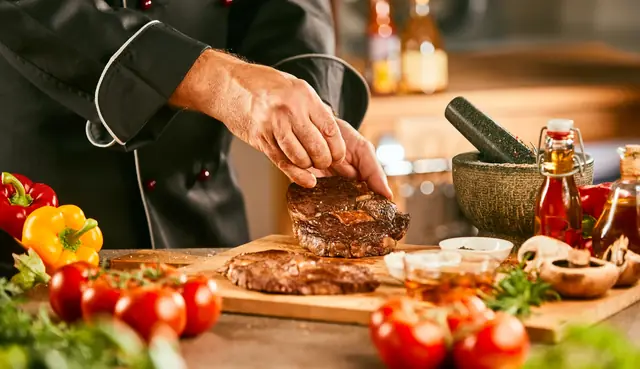 Koch würzt ein gegrilltes Stück Fleisch auf einem Holzbrett, daneben Tomaten und Paprika